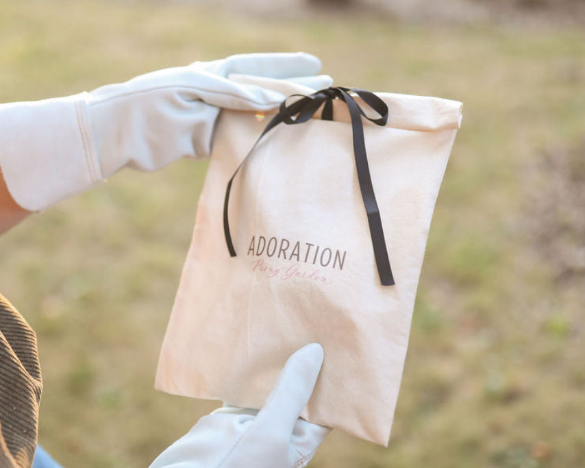 A person wearing white gloves holds a beige fabric pouch with a black ribbon and the word 'ADORATION' printed on it, photographed outdoors with a blurred grassy background.