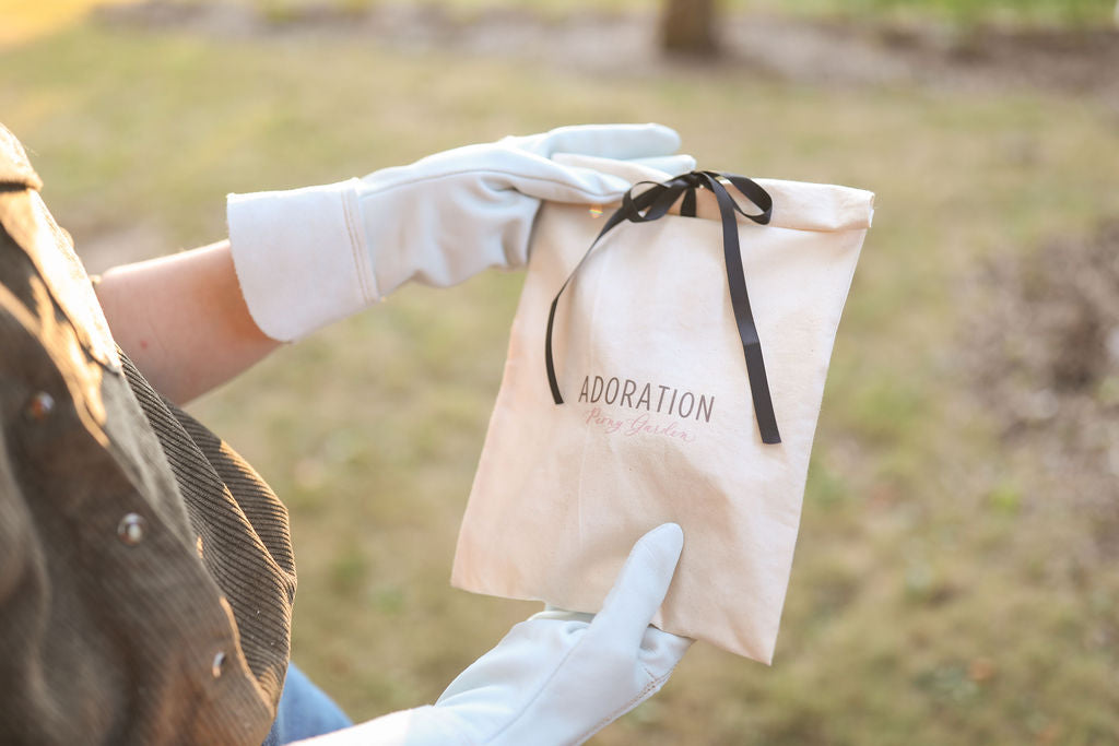 A person wearing white gloves holds a beige fabric pouch with a black ribbon and the word 'ADORATION' printed on it, photographed outdoors with a blurred grassy background.