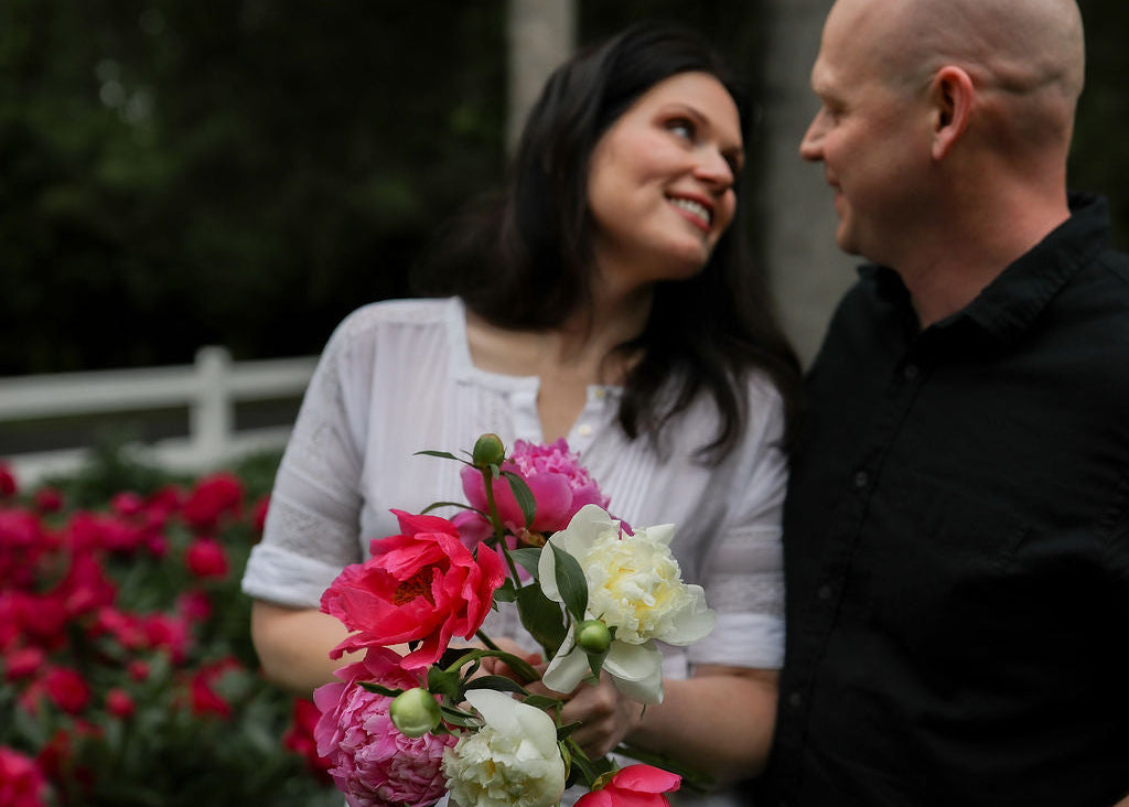 A man and a woman in a white dress holding peony flowers in a garden setting.